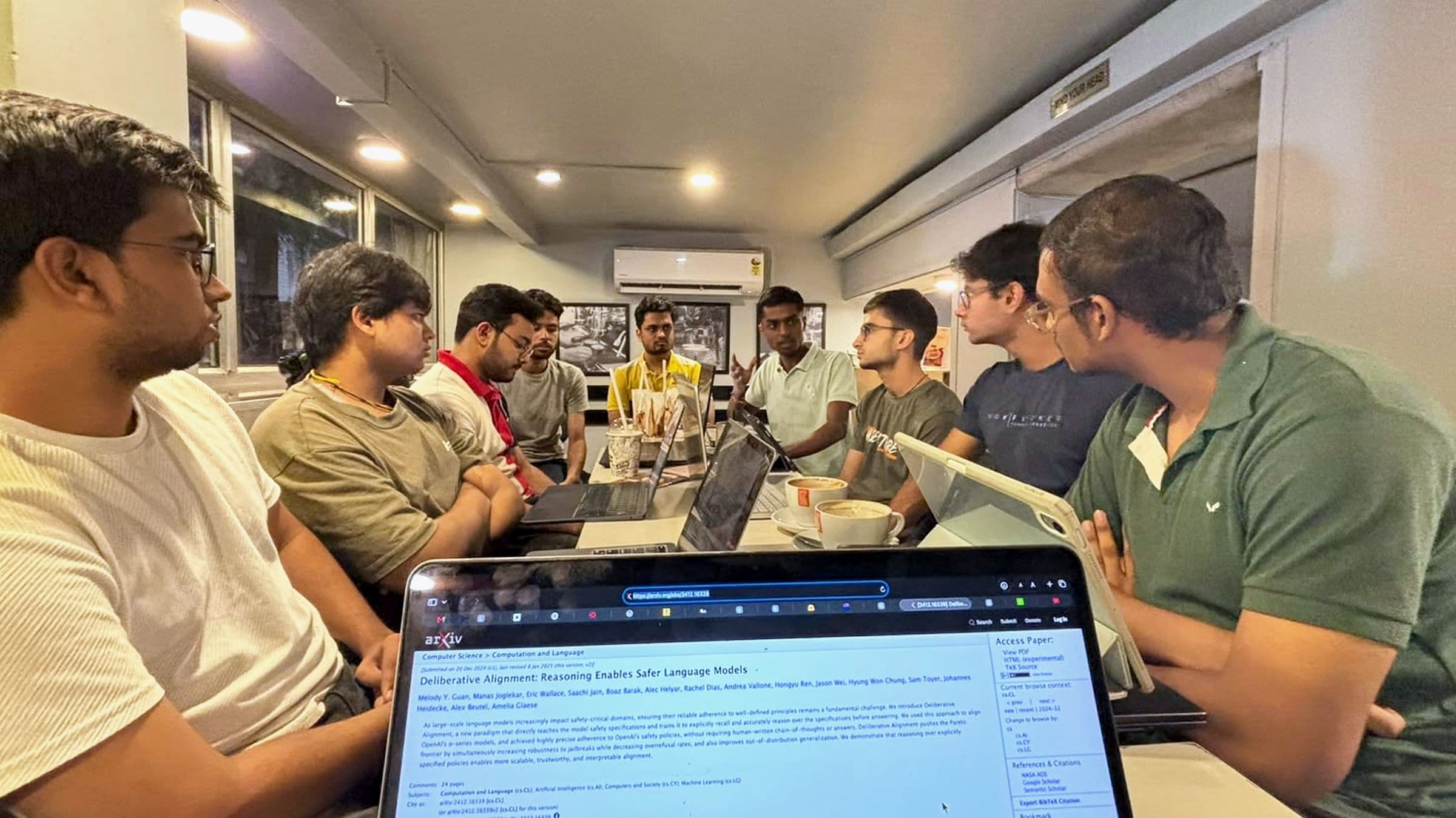 Participants around a long café table with laptops and coffee, listening as one of them makes a point